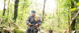 Vater und Sohn sitzen bei einem Vater & Sohn Wildniscamp auf einem umgekippten Baumstamm und schnitzen zusammen mit einem Messer ein Stück Holz.