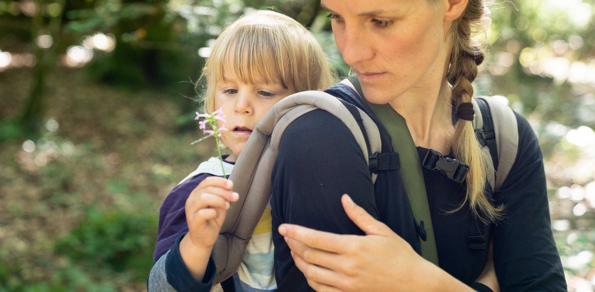 Bei unserer Wildnispädagogik Familien Aktivität bei Berlin sitzt ein Kind auf dem Rücken seiner Mutter und betrachtet staunend eine Blume.