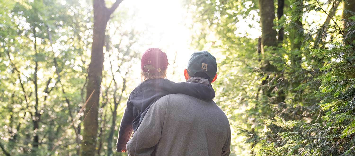 Ein Vater steht bei unserer Wildnispädagogik Familien Aktivität in Berlin mitten im Wald und hat seinen Sohn im Arm. Beide schauen zum Sonnenaufgang.