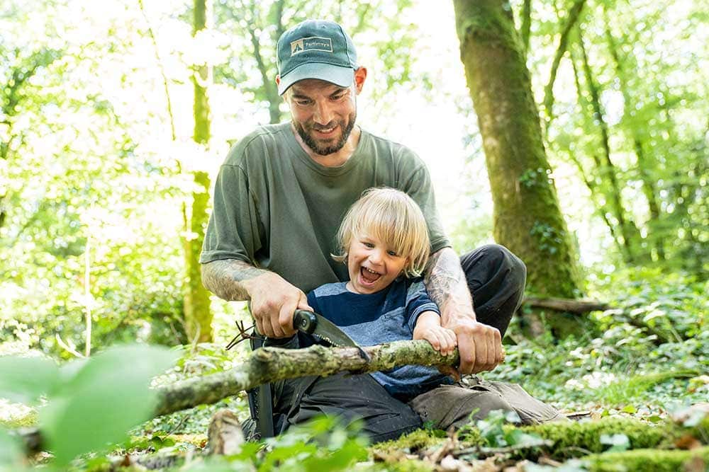 Vater und Sohn sägen gemeinsam, bei unserer Wildnispädagogik Vater & Kind Wildnis Kurs bei Berlin, einen Ast mit einer Säge durch.