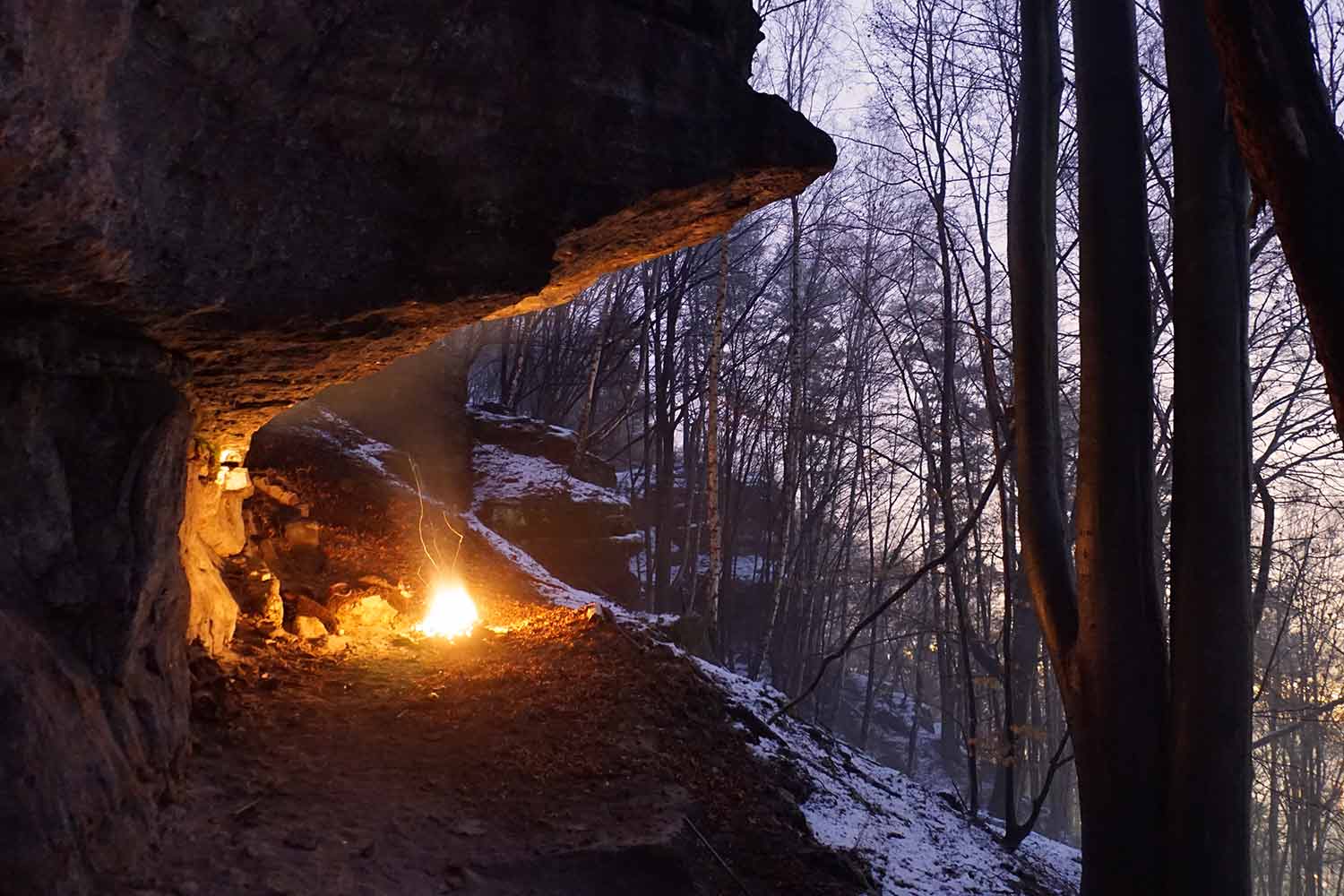 Eine Landschaftsaufnahme unseres Seminarorten für die Wildnispädagogik. In einer Felskante ist ein Feuer entzündest. Im Hintergrund liegt Schnee an den Berghängen.