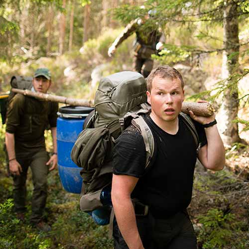 3-tage-survival-training-wochenende Drei Schüler des Survival-Trainings, tragen mit einer improvisierten Trage aus Holz eine Lebensmittel Tonne durch den Wald. Sie Wollen mitten in er Natur eine Survival-Camp errichten.