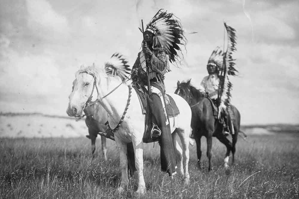 Ein historisches Foto mit drei nordamerikanischen Indigenen auf Pferden in einer weiten Landschaft.