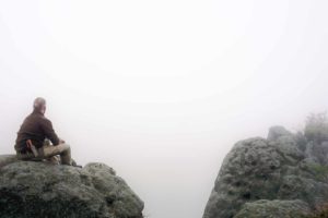 Ein Mann sitzt bei seinem Survival und Bushcraft-Training auf einem Felsen und schau in den Nebel. Unser Survival-Psychologie Kurs mit seine Resilienz-Training und Mentales-Training bereitet die Teilenehmer auf das Leben in der Wildnis vor.