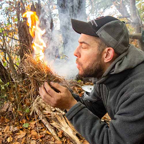 Der Survival-Experte Maurice Ressel zeigt den Teilnehmern wie er mit dem Bow-Drill ein Feuer entfachen kann. Er hält gerade den Zunder in der Hand und pustet hinein. Eine große Flamme ist zu sehen.