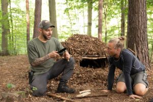 Im Holistic-Survival-Kurs zeigt der Mentor Maurice Ressel einem Teilnehmer neben einer Laubhütte den Bau eins Bow-Drills.