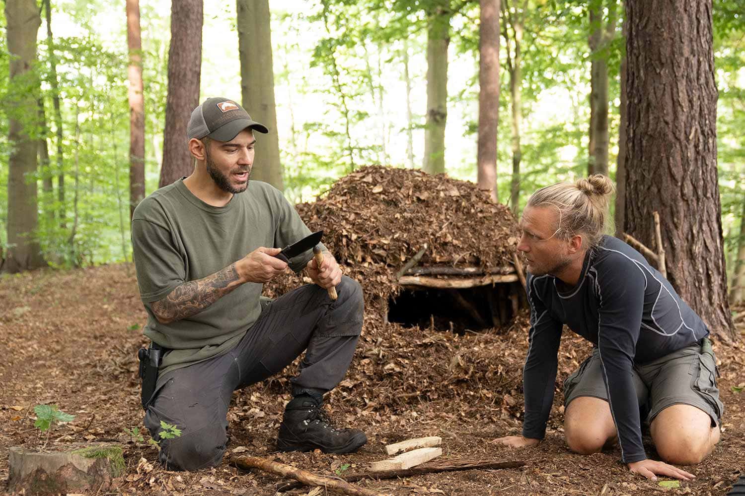 Im Holistic-Survival Kurs zeigt der Mentor Maurice Ressel einem Teilnehmer neben einer Laubhütte den Bau eins Bow-Drills.