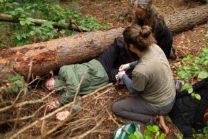 Eine Schauspielerin spielt in einem Erste-Hilfe Szenario eine Stichverletzung durch einen stumpfen Gegenstand. Sie liegt auf dem Waldboden neben einen umgekippten Baum und zwei Teilnehmer des Erste-Hilfe-Kurses legen der Person einen Druckverband an. Das ganze Szenario findet Outdoor in einem Waldgebiet statt.