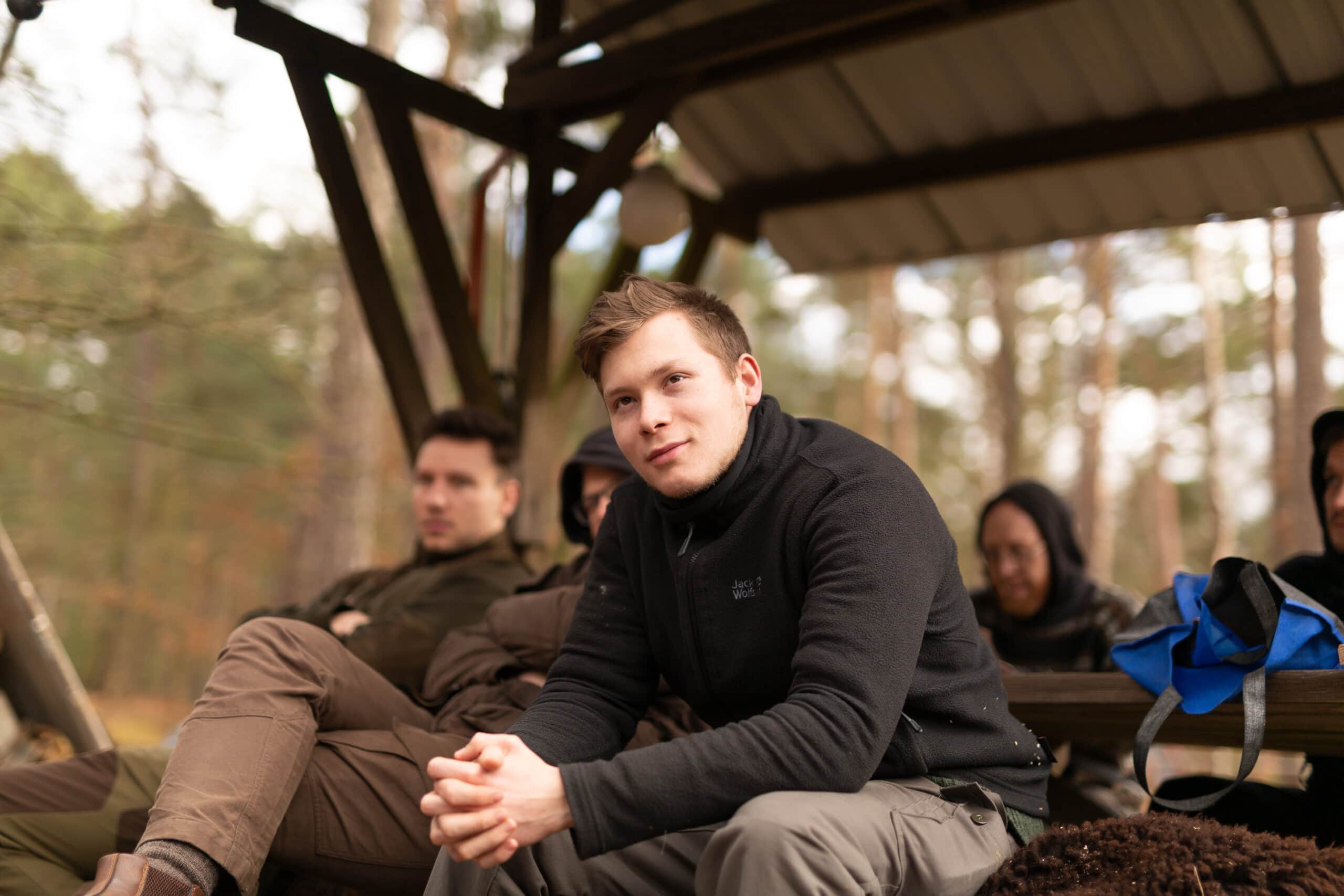 Jagdschüler sitzen auf Holzbänken an der Lagerfeuerstelle der Jagdschule Lupus. Sie hören dem Jagd-Experten Kai Hagemann aufmerksam zu.