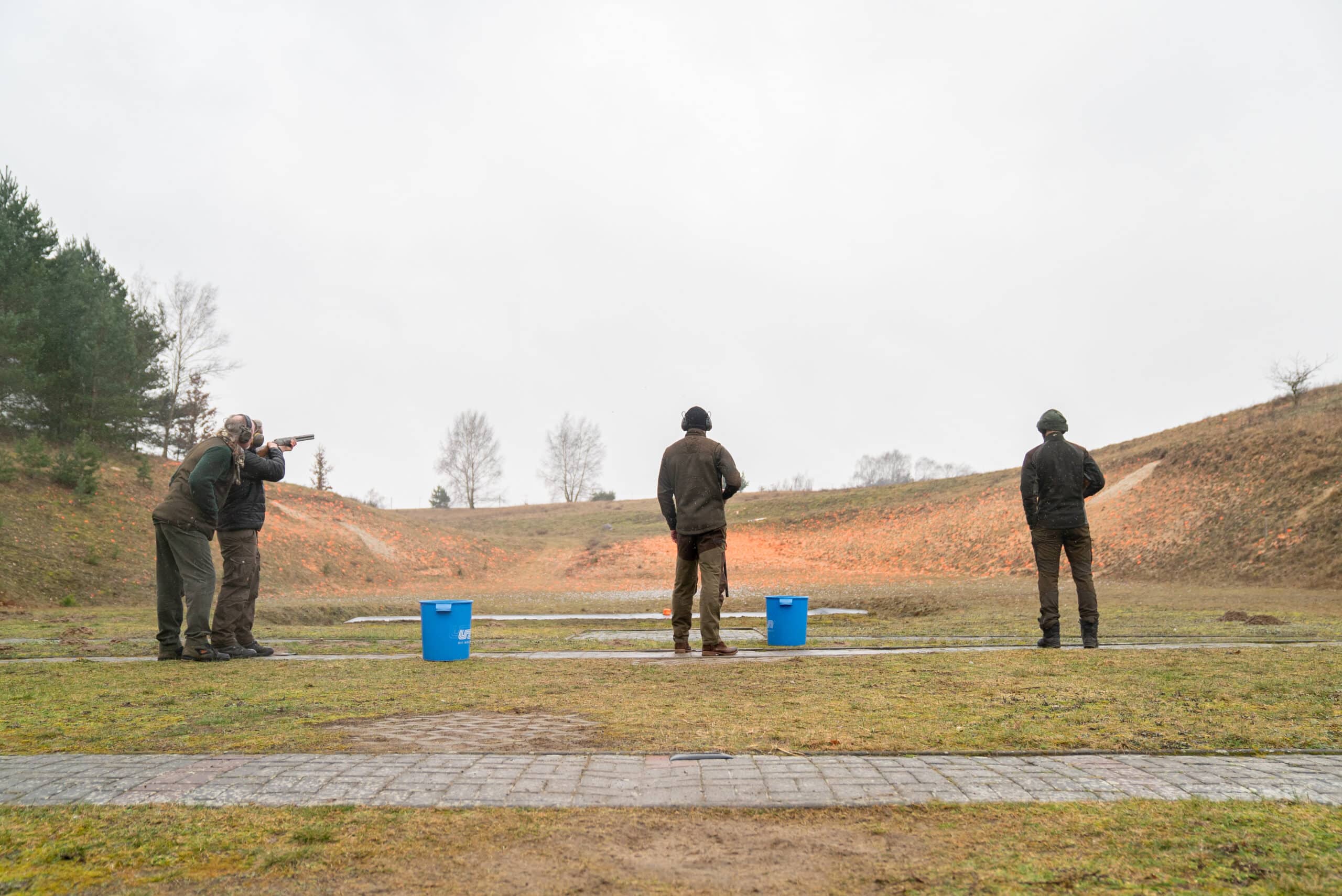 Drei Jagdschüler stehen in der Reihe am Trap Schießstand der Jagdschule. Der Line Schüler wird gerade von Schießtrainer Mike Gellenbeck betreut.